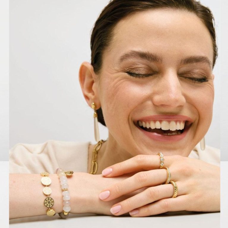 Schmuck für Damen Frau mit braunem Haar, einem weißen Blusenshirt und goldener Schmuck sitzt entspannt.